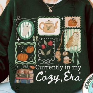 May include: A forest green crewneck sweatshirt featuring a graphic print of various items including a ghost, teapot, pumpkins, an owl, books, and a broom. The text reads, "Currently in my Cozy Era." The design is arranged in a grid-like pattern with decorative borders and bows.