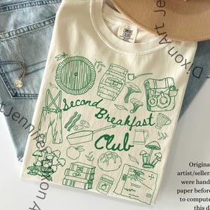 May include: Cream-coloured t-shirt with a green doodle design featuring the text "Second Breakfast Club." The design includes illustrations of a hobbit hole, food, and other related imagery. The shirt is paired with blue jeans and a beige hat.