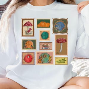 May include: White sweatshirt featuring a collection of whimsical postage stamp designs. The stamps depict mushrooms, pumpkins, and fairy doors, with various denominations. The overall theme is fantasy and nature.