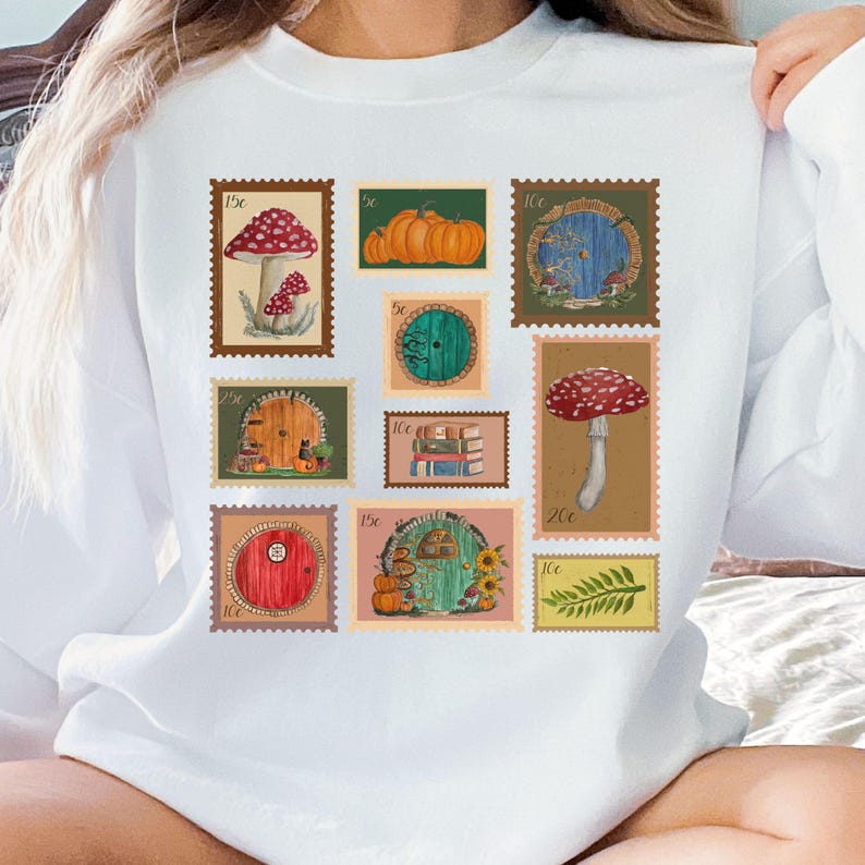 May include: White sweatshirt featuring a collection of whimsical postage stamp designs. The stamps depict mushrooms, pumpkins, and fairy doors, with various denominations. The overall theme is fantasy and nature.