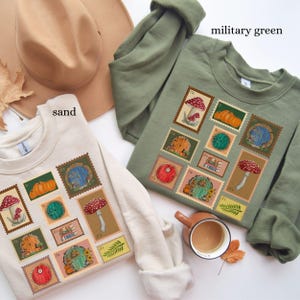 May include: Sand-colored and military green sweatshirts with a whimsical stamp design featuring mushrooms, pumpkins, and hobbit-hole doors. A brown hat and a cup of coffee are also in the image. The sweatshirts are made of a soft material.