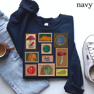 May include: Navy blue sweatshirt featuring a collection of whimsical postage stamp designs. The stamps depict mushrooms, pumpkins, and hobbit door illustrations. The word "navy" is printed in the upper right corner.