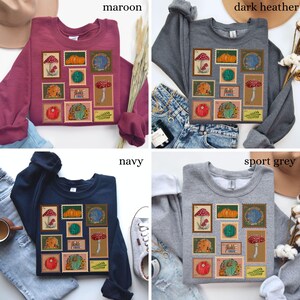 May include: Four sweatshirts in maroon, dark heather, navy, and sport grey. Each sweatshirt features a collection of illustrated stamps with mushrooms, pumpkins, and whimsical doors. The word "maroon", "dark heather", "navy", and "sport grey" are printed on the image.