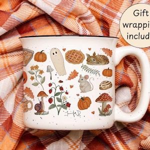 May include: White ceramic mug with a black rim, decorated with autumn-themed illustrations. The design includes pumpkins, a ghost, pie, a deer, a mouse, mushrooms, and leaves. The mug is set against a plaid fabric background. The text "Gift wrapping included" is also visible.