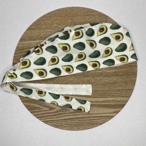 May include: White surgical scrub hat with a repeating pattern of green avocados on a white background. The hat has a long tie that can be tied at the back of the head.