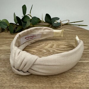May include: A beige knotted headband made of linen fabric. The headband has a label that says "emintees' Craft Room".