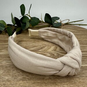 May include: A beige knotted headband made of fabric. The headband is on a wooden surface.