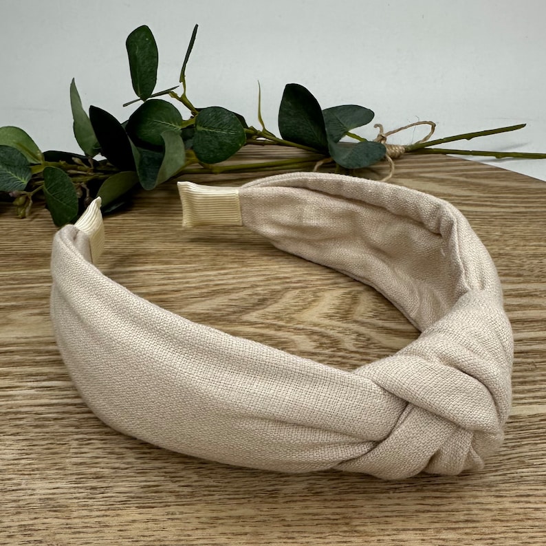 May include: A beige knotted headband made of fabric. The headband is on a wooden surface.