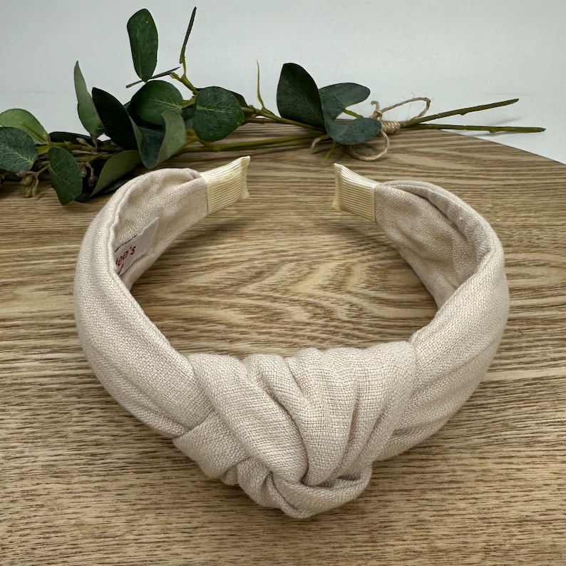 May include: A beige knotted headband made of linen fabric. The headband has a simple design and is perfect for adding a touch of style to any outfit.