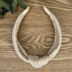May include: A beige knotted headband made of fabric. The headband is on a wooden surface.