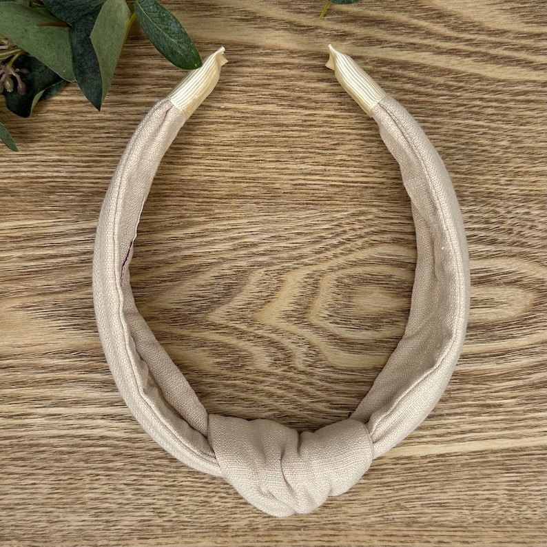 May include: A beige knotted headband made of fabric. The headband is on a wooden surface.