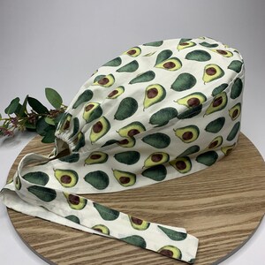 May include: White scrub hat with a green avocado print. The hat has a tie that is also printed with avocados.