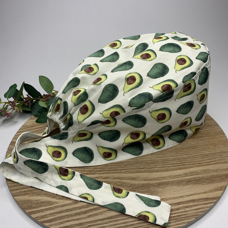 May include: White scrub hat with a green avocado print. The hat has a tie that is also printed with avocados.
