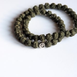 Könnte beinhalten: Ein kreisförmiges Armband aus olivgrünem, geknotetem Kordelmaterial mit einer kleinen, runden, silberfarbenen Metallperle, die ein eingeprägtes Kreuzsymbol aufweist. Die Kordel ist mit einer Reihe gleichmäßiger Knoten in einer Schlaufe versehen.