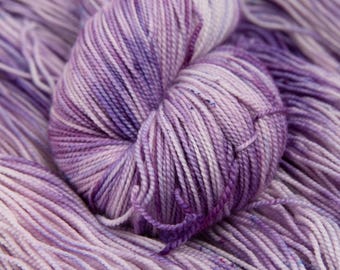 May include: A close-up of a skein of yarn in shades of purple, lavender, and white. The yarn is tightly wound, with visible strands and a soft, textured appearance. The color variations create a visually appealing, handcrafted look.