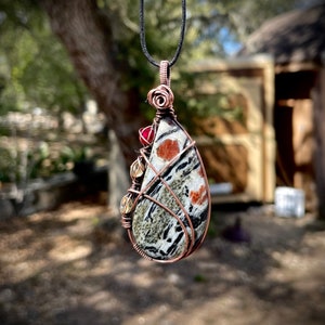 Op de afbeelding: Een handgemaakte hanger met een druppelvormige steen, met een kenmerkend patroon van zwarte, witte en oranje markeringen. De steen is omwikkeld met koperdraad en versierd met rode en gele kralen, hangend aan een zwart koord.