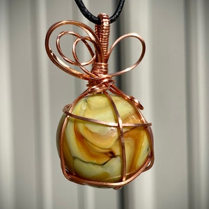 Puede incluir: Un collar con colgante artesanal que presenta una piedra esférica veteada en tonos amarillos y marrones, encerrada en una jaula de alambre de cobre. El colgante cuelga de un cordón negro, mostrando el intrincado trabajo de alambre y la piedra natural.