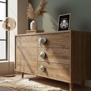 Puede incluir: Un aparador de madera con tres cajones y una puerta, con tiradores en forma de animales. El aparador está coronado con un jarrón de flores secas y una ilustración enmarcada. Una alfombra con estampado de cebra está en el suelo.