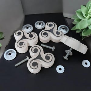 Könnte beinhalten: Vier beigefarbene Schrankknöpfe mit einem schwarzen Wirbelmuster. Jeder Knopf hat eine silberne Schraube und eine silberne Unterlegscheibe.