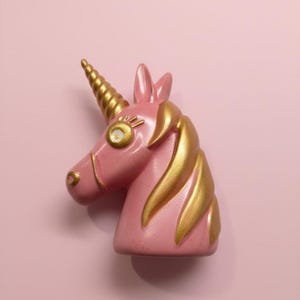Puede incluir: Una figura de cabeza de unicornio rosa con cuerno y melena dorados. El unicornio tiene un diseño caprichoso con un cuerno y una melena dorados detallados, y un cuerpo rosa claro. El fondo es rosa claro liso.