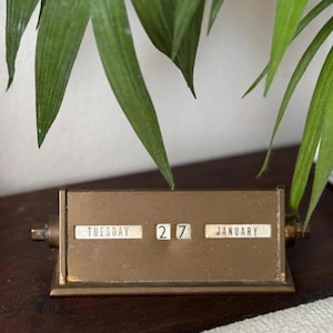 May include: A vintage brass perpetual calendar displaying "Tuesday 27 January". The calendar has a rectangular base with rotating date and month indicators. Green leafy plant in the background.