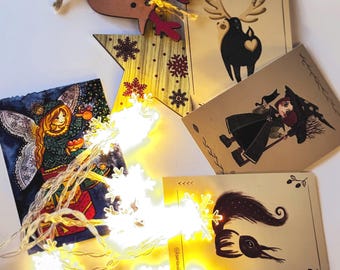 May include: A collection of Christmas-themed decorations and cards. Includes a wooden reindeer ornament, a star-shaped ornament with snowflake designs, and several illustrated cards featuring reindeer, a witch, and a whimsical creature. A string of snowflake lights adds a festive touch.