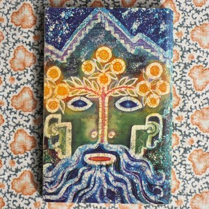 K&ouml;nnte beinhalten: Ein Hardcover-Buch mit einer lebendigen, abstrakten Illustration. Das Cover zeigt ein gesichts&auml;hnliches Design mit einer baumartigen Struktur, einem blauen Bart und einer Bergkette oben. Die Farbpalette umfasst Blau-, Gr&uuml;n- und Oranget&ouml;ne.