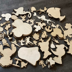 May include: A collection of unfinished wooden cutouts in various shapes, including cartoon characters, castle, and Mickey Mouse silhouettes. The cutouts are made of light-colored wood and are scattered on a dark surface.