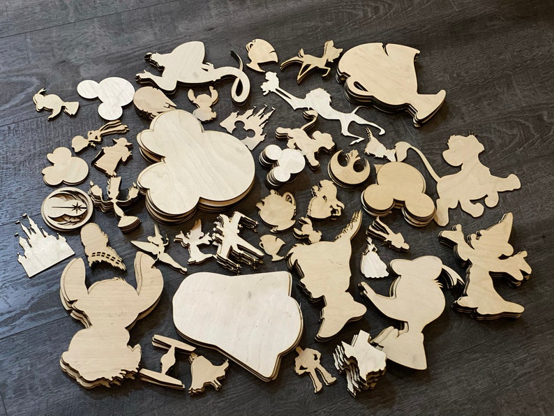 May include: A collection of unfinished wooden cutouts in various shapes, including cartoon characters, castle, and Mickey Mouse silhouettes. The cutouts are made of light-colored wood and are scattered on a dark surface.