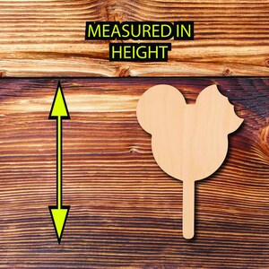 May include: A light-colored wooden Mickey Mouse-shaped cutout on a stick, with a bite taken out of the ear. The text "MEASURED IN HEIGHT" is above the cutout. The background is a dark brown wooden surface.