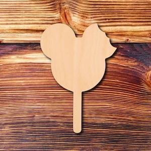 May include: A wooden Mickey Mouse-shaped cutout on a stick, perfect for crafts. The cutout is light brown and features the iconic mouse ears and a bow. The background is a dark brown wooden surface.