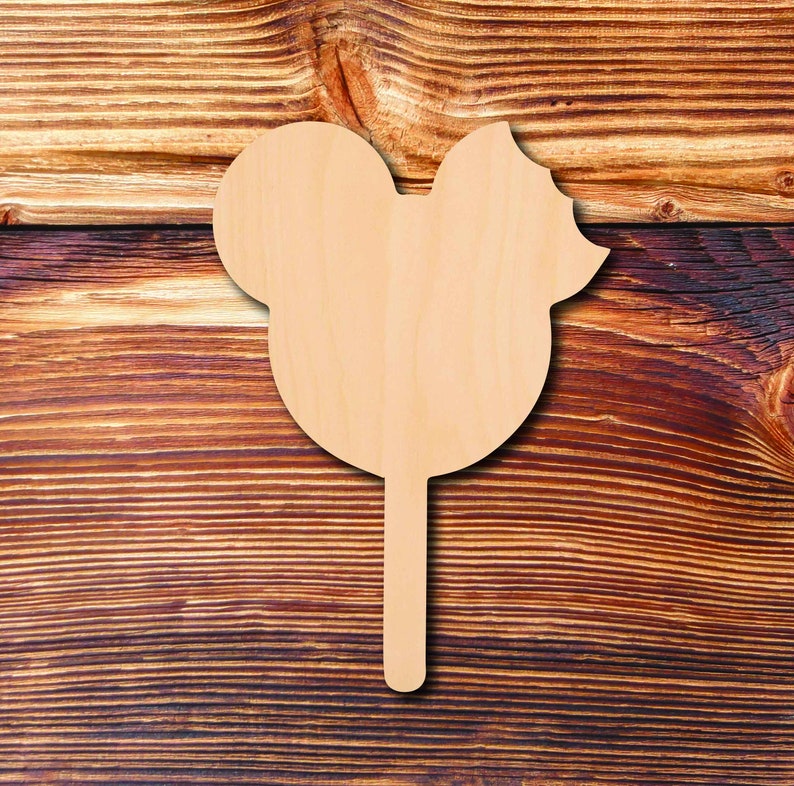 May include: A wooden Mickey Mouse-shaped cutout on a stick, perfect for crafts. The cutout is light brown and features the iconic mouse ears and a bow. The background is a dark brown wooden surface.