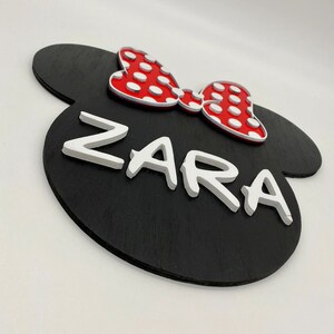 May include: Black wooden sign shaped like Mickey Mouse ears with a red bow with white polka dots and the name "ZARA" in white raised letters.
