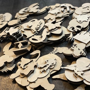 May include: A collection of laser-cut wooden shapes, including various cartoon characters and castle silhouettes. The shapes are stacked and scattered, showcasing the natural wood grain and intricate details of the designs. The items are unfinished and ready for crafting.