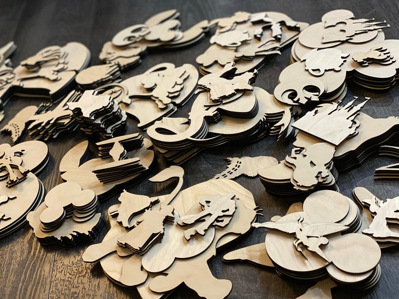 May include: A collection of laser-cut wooden shapes, including various cartoon characters and castle silhouettes. The shapes are stacked and scattered, showcasing the natural wood grain and intricate details of the designs. The items are unfinished and ready for crafting.