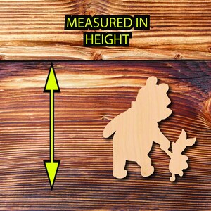 May include: Wooden cutout of Winnie the Pooh and Piglet holding hands. The cutout is on a brown wood background.  MEASURED IN HEIGHT is written above the cutout.