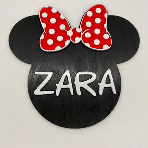 May include: Black wooden cutout of a Mickey Mouse head with a red bow with white polka dots and the name "ZARA" written in white letters.