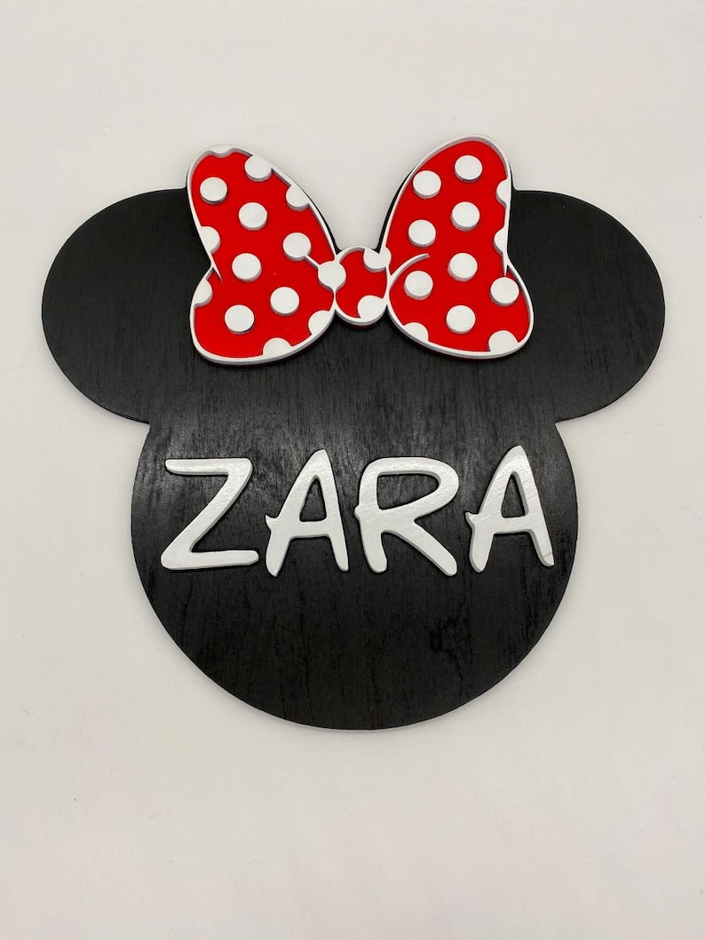 May include: Black wooden cutout of a Mickey Mouse head with a red bow with white polka dots and the name "ZARA" written in white letters.