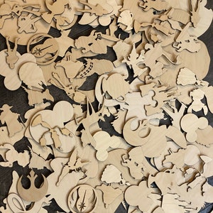 May include: A collection of unfinished wooden cutouts in various shapes and sizes, including character silhouettes and iconic symbols. The cutouts are made of light-colored wood and are scattered across a dark surface. The shapes include Mickey Mouse, Star Wars symbols, and other cartoon characters.