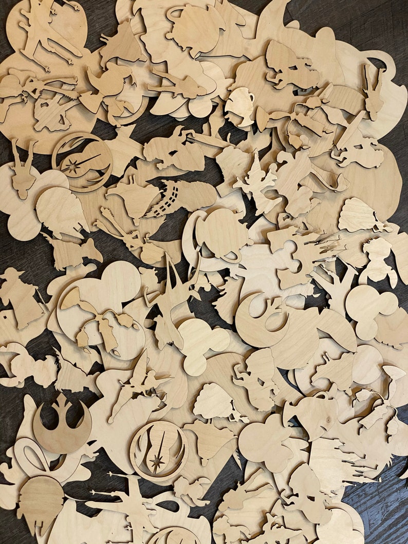May include: A collection of unfinished wooden cutouts in various shapes and sizes, including character silhouettes and iconic symbols. The cutouts are made of light-colored wood and are scattered across a dark surface. The shapes include Mickey Mouse, Star Wars symbols, and other cartoon characters.