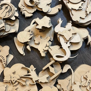 May include: A collection of laser-cut wooden shapes, including various cartoon characters and Star Wars-themed designs. The shapes are unpainted and feature intricate details, suggesting they are intended for crafting or decoration. The pieces are scattered across a dark surface.