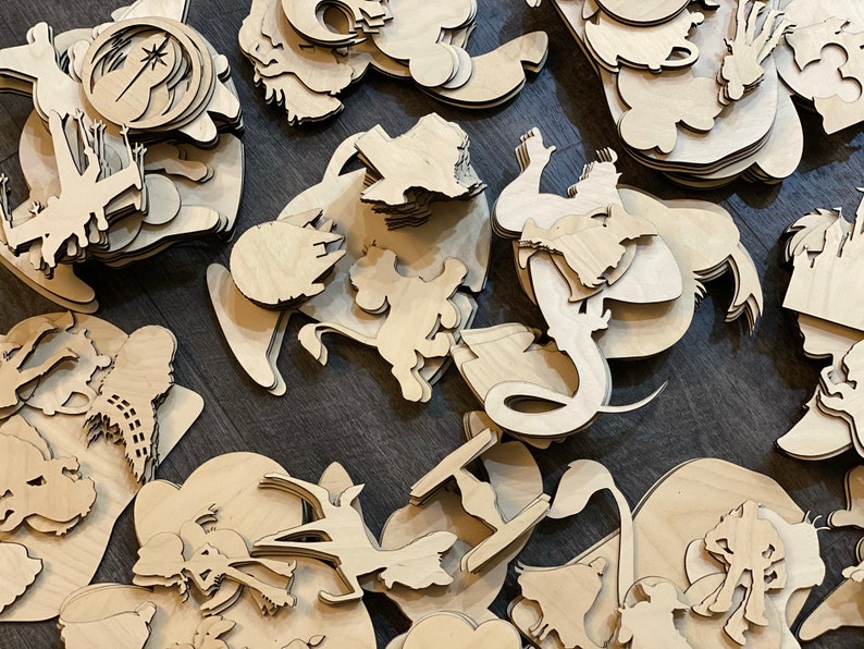 May include: A collection of laser-cut wooden shapes, including various cartoon characters and Star Wars-themed designs. The shapes are unpainted and feature intricate details, suggesting they are intended for crafting or decoration. The pieces are scattered across a dark surface.