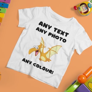 May include: White t-shirt with a cartoon pterodactyl design. The text "ANY TEXT ANY PHOTO ANY COLOUR!" is printed on the shirt.