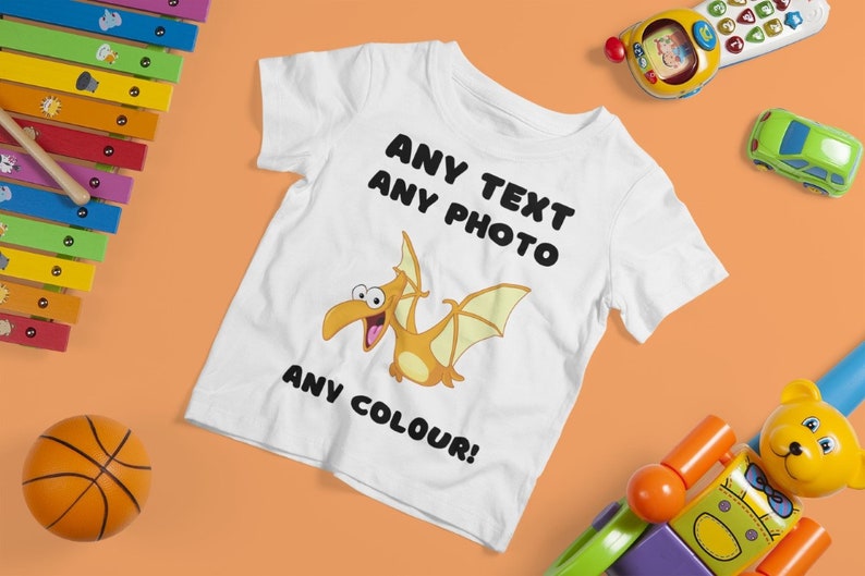 May include: White t-shirt with a cartoon pterodactyl design. The text "ANY TEXT ANY PHOTO ANY COLOUR!" is printed on the shirt.