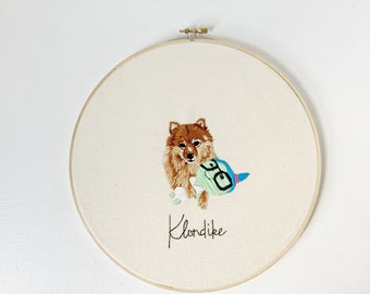 May include: Embroidered hoop art featuring a brown and white dog with a green and blue toy. The dog's name, "Klondike", is embroidered below the image.