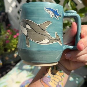 May include: A light blue ceramic mug featuring a hand-painted illustration of a large black and white orca whale swimming across the center, with a smaller blue and white shark depicted above it. The mug has a sturdy handle and a smooth glazed finish.
