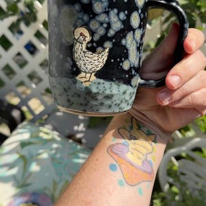 May include: A ceramic mug with a black glaze featuring a speckled pattern of light blue and white spots. The mug displays a hand-drawn illustration of a chicken with a red comb on the front. It has a curved handle and a textured base.