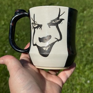 May include: A ceramic mug with a black handle and a design featuring a stylized face. The face is depicted in black on a white background, with a black stripe on the side. The mug is held in a hand.
