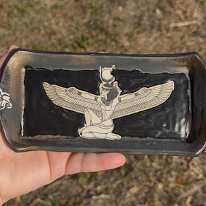 May include: A rectangular ceramic tray with a black interior and a gray and gold rim. The tray features a cream-colored illustration of a winged deity. The tray has handles with decorative designs on each end.