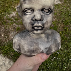 May include: A ceramic planter shaped like a baby's head and torso. The planter is primarily gray with black shading and a red interior. The face has a somber expression with closed eyes and a slightly open mouth. The planter is held in a hand.
